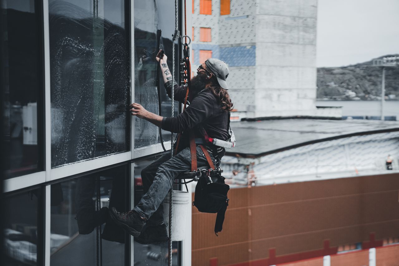 Un homme nettoie les vitres d’un gratte-ciel avec un harnais, illustrant le travail urbain en hauteur.